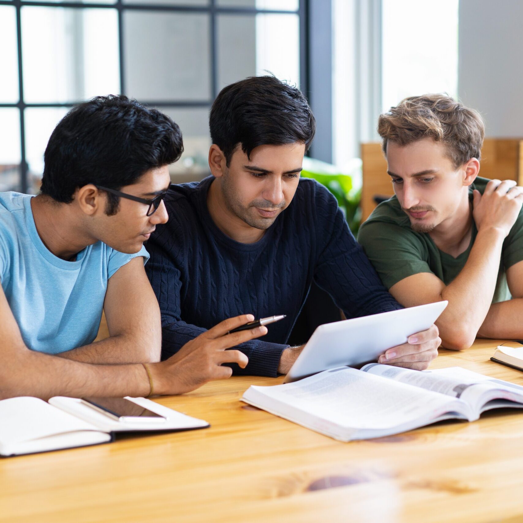 Focused students using tablet and discussing information. Young men studying and sitting at desk in classroom or library. Education and technology concept.